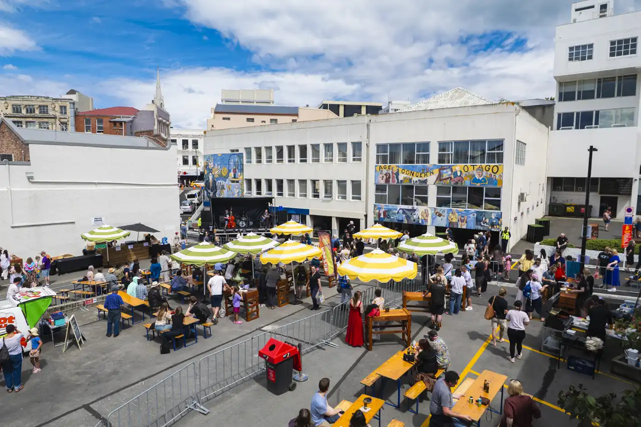 A wide shot of the Beats and Eats event on Bath Street, which shows people standing and sitting at tables and under colourful sun umbrellas, enjoying food, drink and a live band playing the double bass, drums and electric guitar