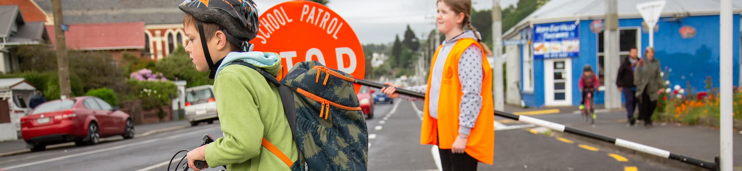 A primary school child walks his bike across the school crossing, while a child on road patrol holds the lollipop stop sign behind them.