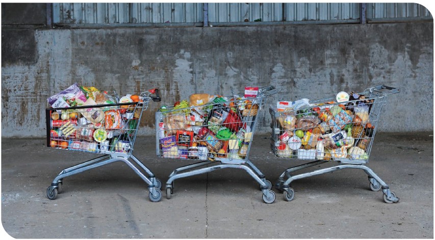 Three shopping carts filled with various groceries are lined up side by side against a concrete wall. The carts contain a variety of items including bread, vegetables, snacks, and packaged goods. This represents the amount of food wasted in the average New Zealand household.